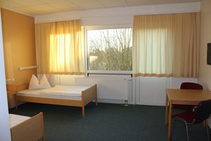 Auf dem Bild sind zwei frischbezogene Betten und eine Sitzgruppe. Das Zimmer leuchtet in warmen Gelbtönen an den Wänden und durch die Gardinen.