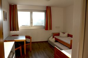 Das Zimmer hat zwei Betten, wovon eins ein Ausziehbett hat. Alle 3 Betten sind frischbezogen. Eine Sitzgruppe steht links neben dem Fenster, welches rot-orange Gardinen hat.