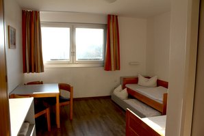 Mehrbettzimmer im Haupthaus Das Zimmer hat zwei Betten, wovon eins ein Ausziehbett hat. Alle 3 Betten sind frischbezogen. Eine Sitzgruppe steht links neben dem Fenster, welches rot-orange Gardinen hat.