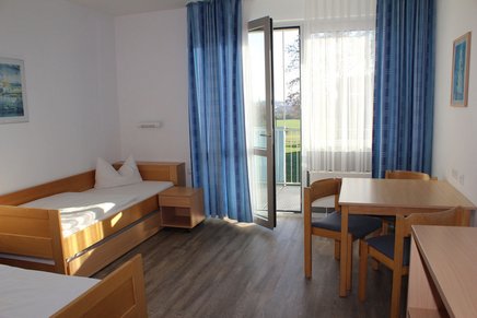 Doppelzimmer im Haupthaus mit Balkonzugang Auf dem Bild sieht man zwei bezogene Betten, eine Sitzgruppe und eine geöffnete Balkontür, die mit blauen Gardinen umrandet ist.