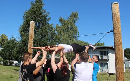 Teambuilding an der Landessportschule Auf dem Bild sieht man Personen, die eine weibliche Person durch das Spinnennetz heben. Das Spinnennetz ist ein Teil des Niedrigseilparcours.