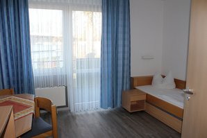 Das Zimmer hat ein frischbezogenes Bett und einen Schreibtisch. Die Gardinen sind erfrischend blau.